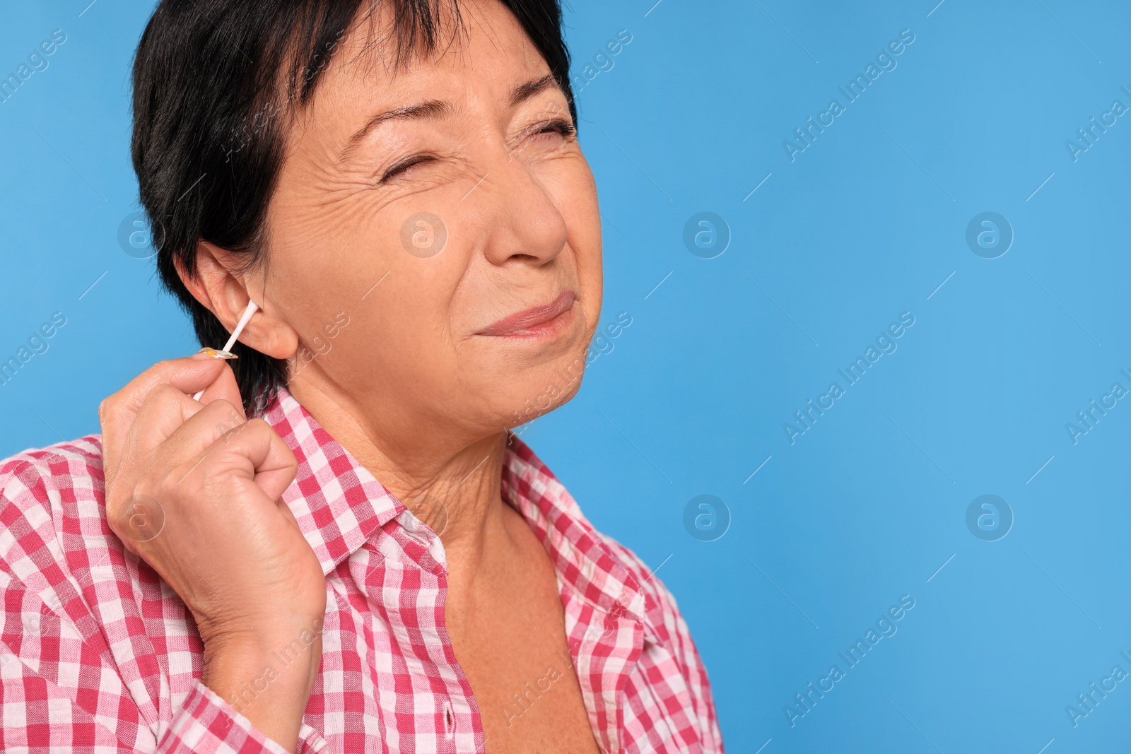 Photo of Senior woman cleaning ear with cotton swab on light blue background, closeup. Space for text