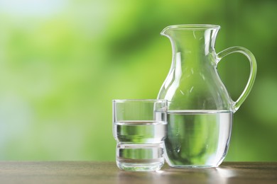 Jug and glass with clear water on wooden table against blurred green background, closeup. Space for text