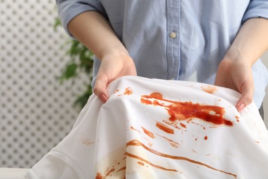 Photo of Woman holding shirt with stain of sauce on blurred background, closeup
