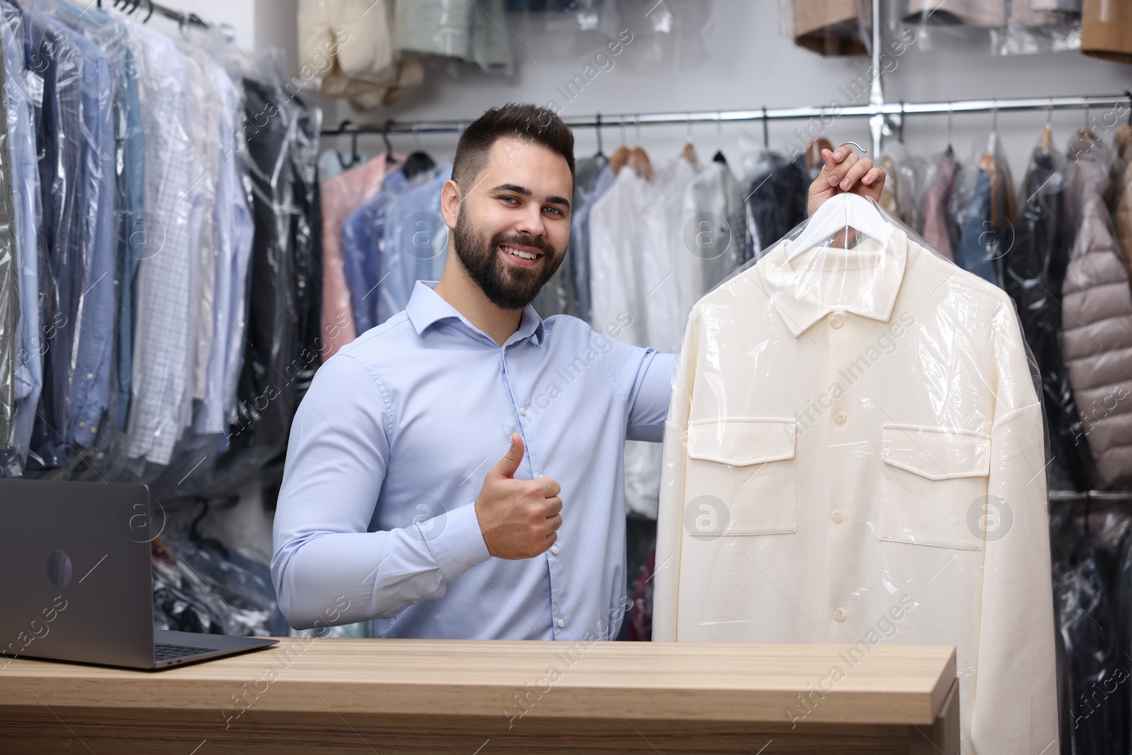 Photo of Dry-cleaning service. Happy worker holding hanger with shirt in plastic bag and showing thumb up at counter indoors