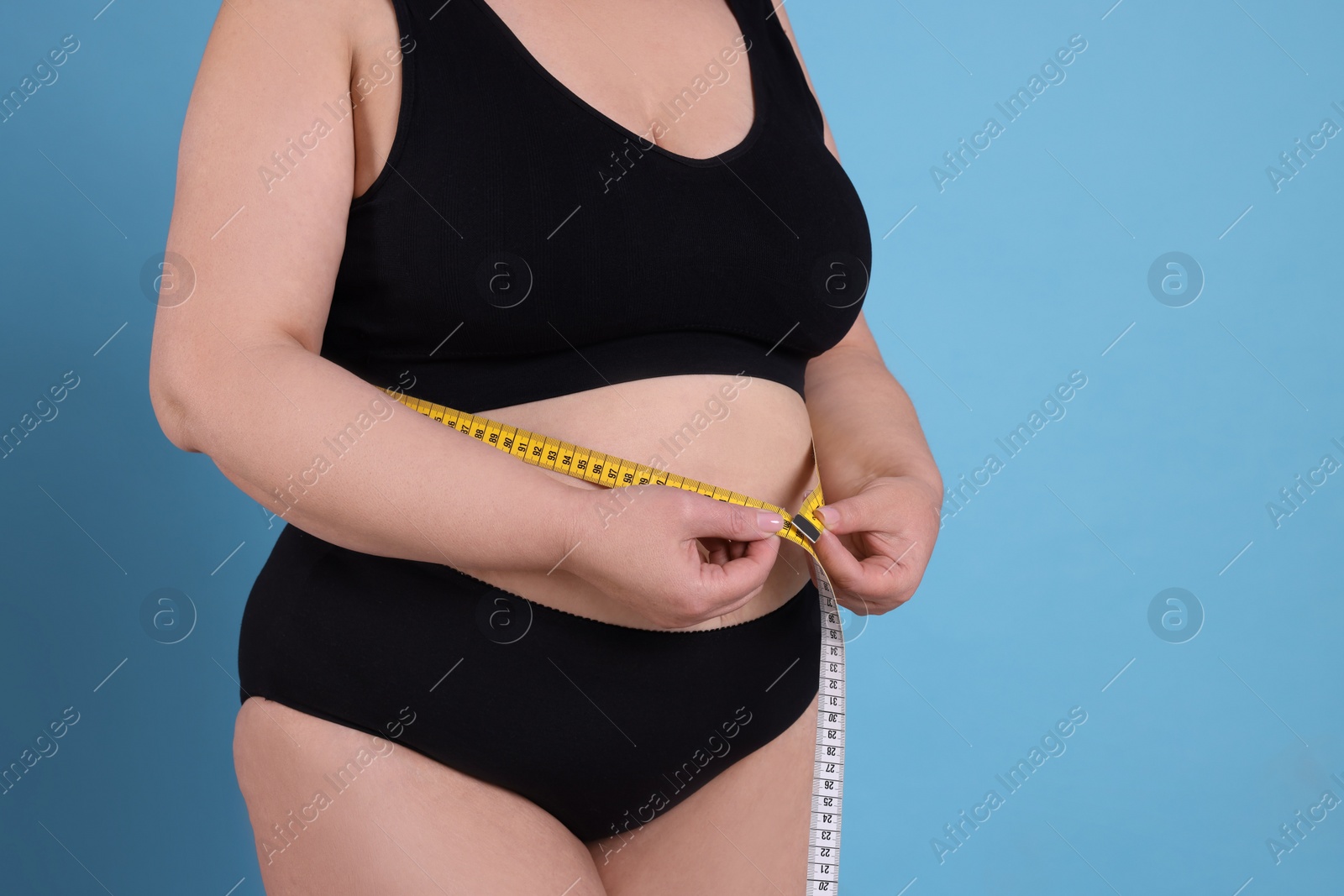 Photo of Overweight woman in underwear measuring waist with tape on light blue background, closeup