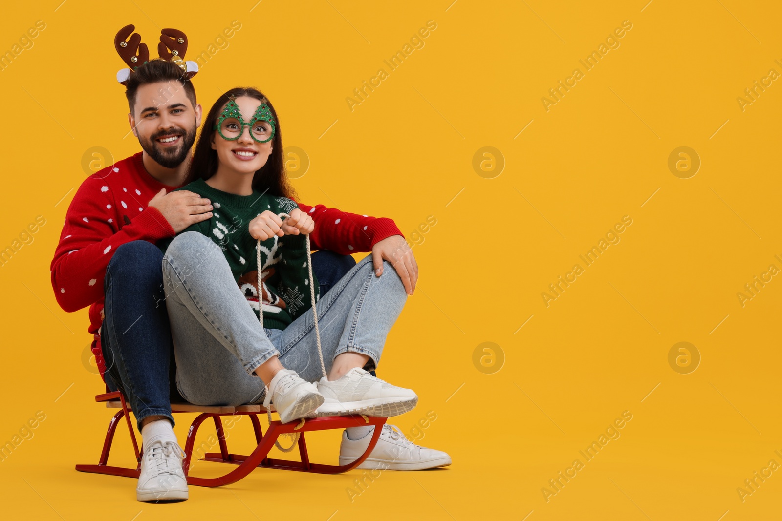 Photo of Happy young couple in Christmas sweaters, reindeer headband and party glasses on sled against orange background. Space for text