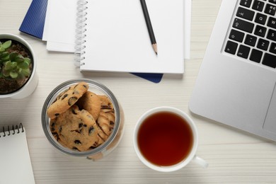 Chocolate chip cookies, cup of tea, office supplies and laptop on white wooden table, flat lay