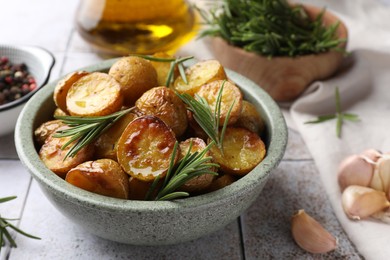 Photo of Bowl with tasty baked potato and aromatic rosemary on white tiled table, closeup