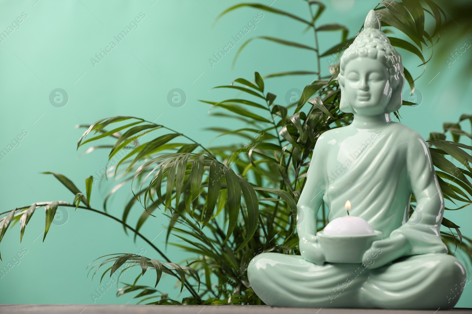 Photo of Buddhism religion. Decorative Buddha statue with burning candle on table and houseplant against turquoise wall