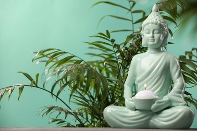 Buddhism religion. Decorative Buddha statue with burning candle on table and houseplant against turquoise wall