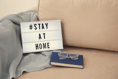 Photo of Book, glasses, plaid and lightbox with hashtag STAY AT HOME on sofa. Message to promote self-isolation during COVID‑19 pandemic