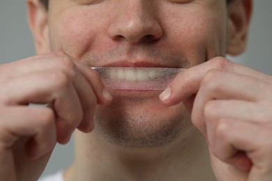 Photo of Young man applying whitening strip on his teeth against light grey background, closeup