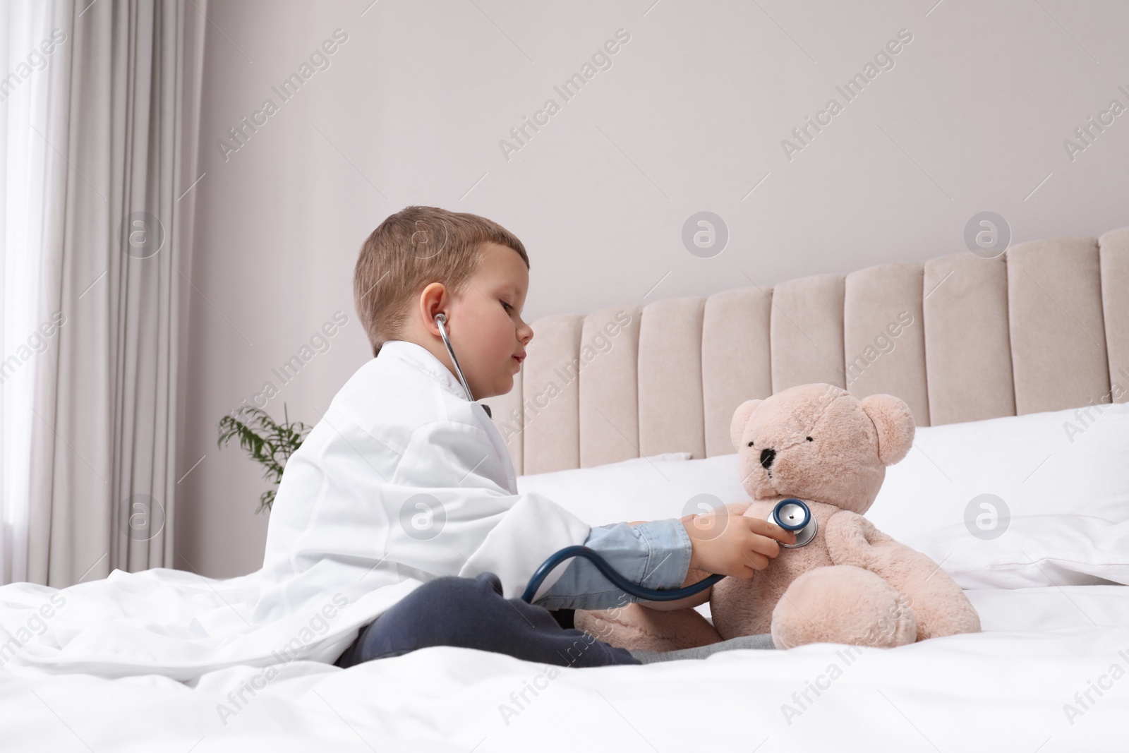 Photo of Cute little boy in pediatrician's uniform playing with stethoscope and toy bear at home