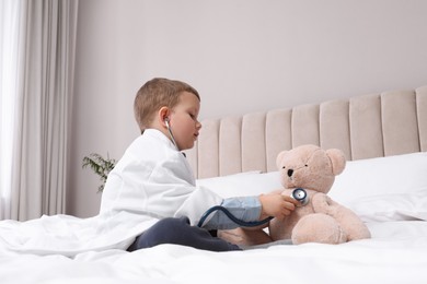Cute little boy in pediatrician's uniform playing with stethoscope and toy bear at home