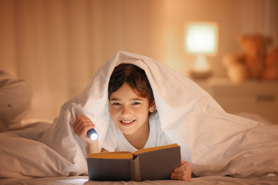 Photo of Boy with flashlight reading book under blanket at home