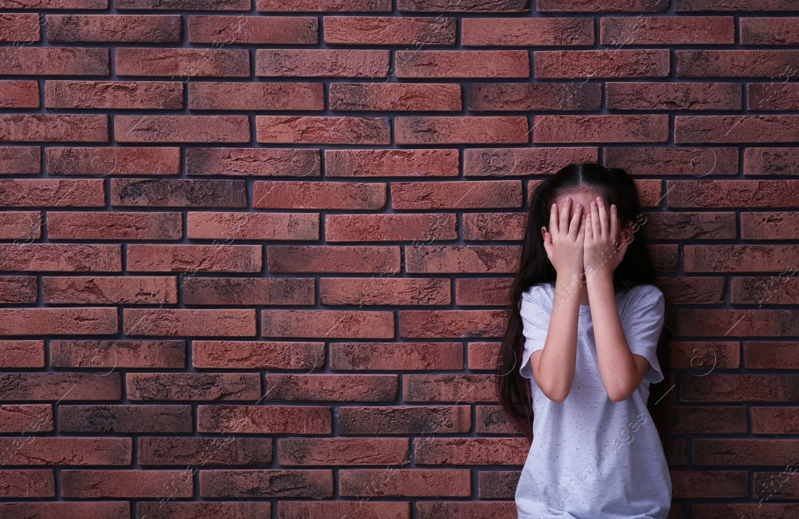 Photo of Sad little girl closing eyes with hands near brick wall, space for text. Child in danger