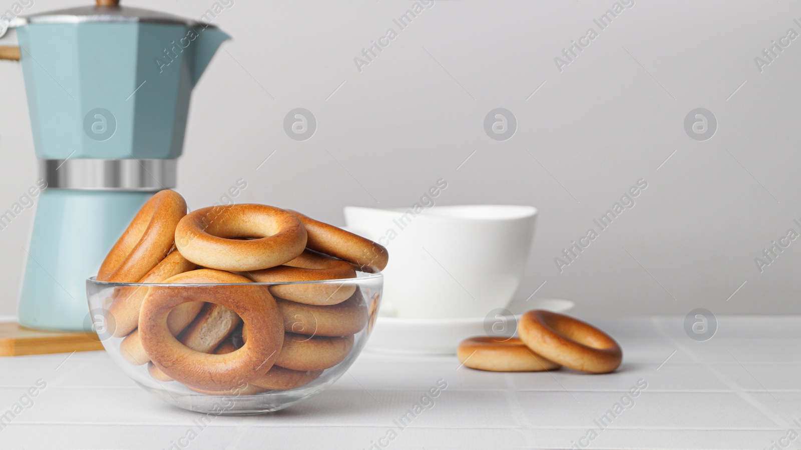 Photo of Bowl with tasty dry bagels (sushki) and cup of hot drink on white tiled table, space for text