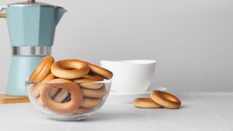 Photo of Bowl with tasty dry bagels (sushki) and cup of hot drink on white tiled table, space for text