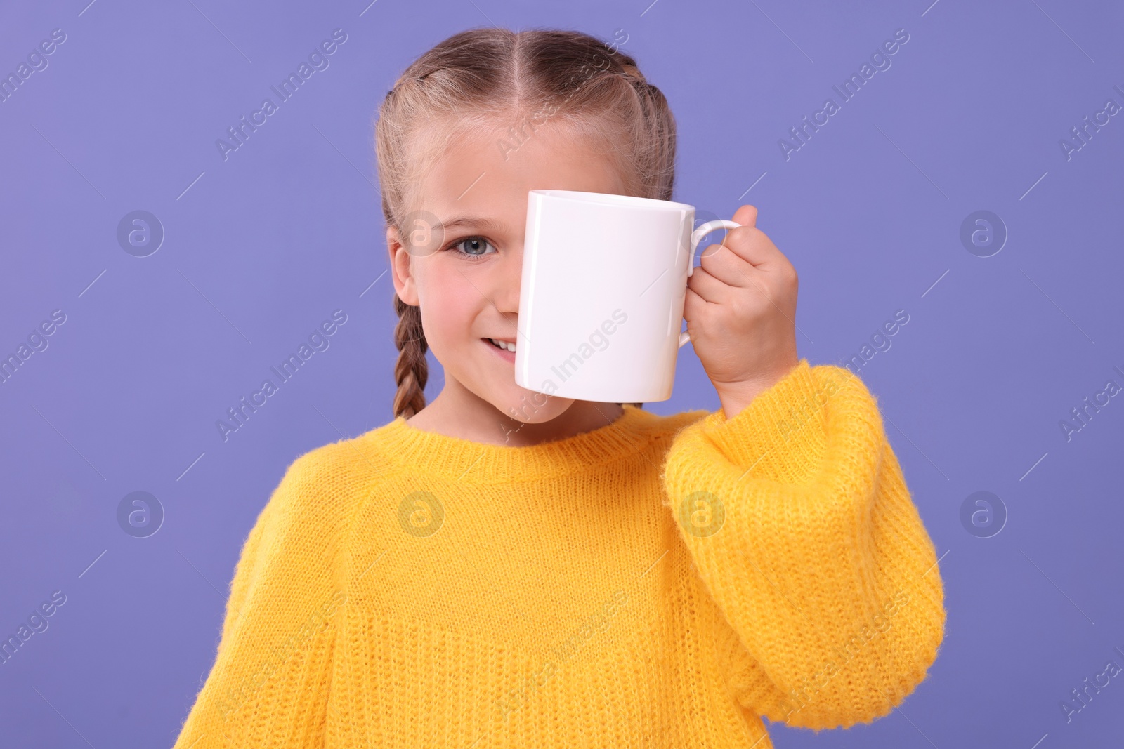 Photo of Happy girl covering eye with white ceramic mug on violet background