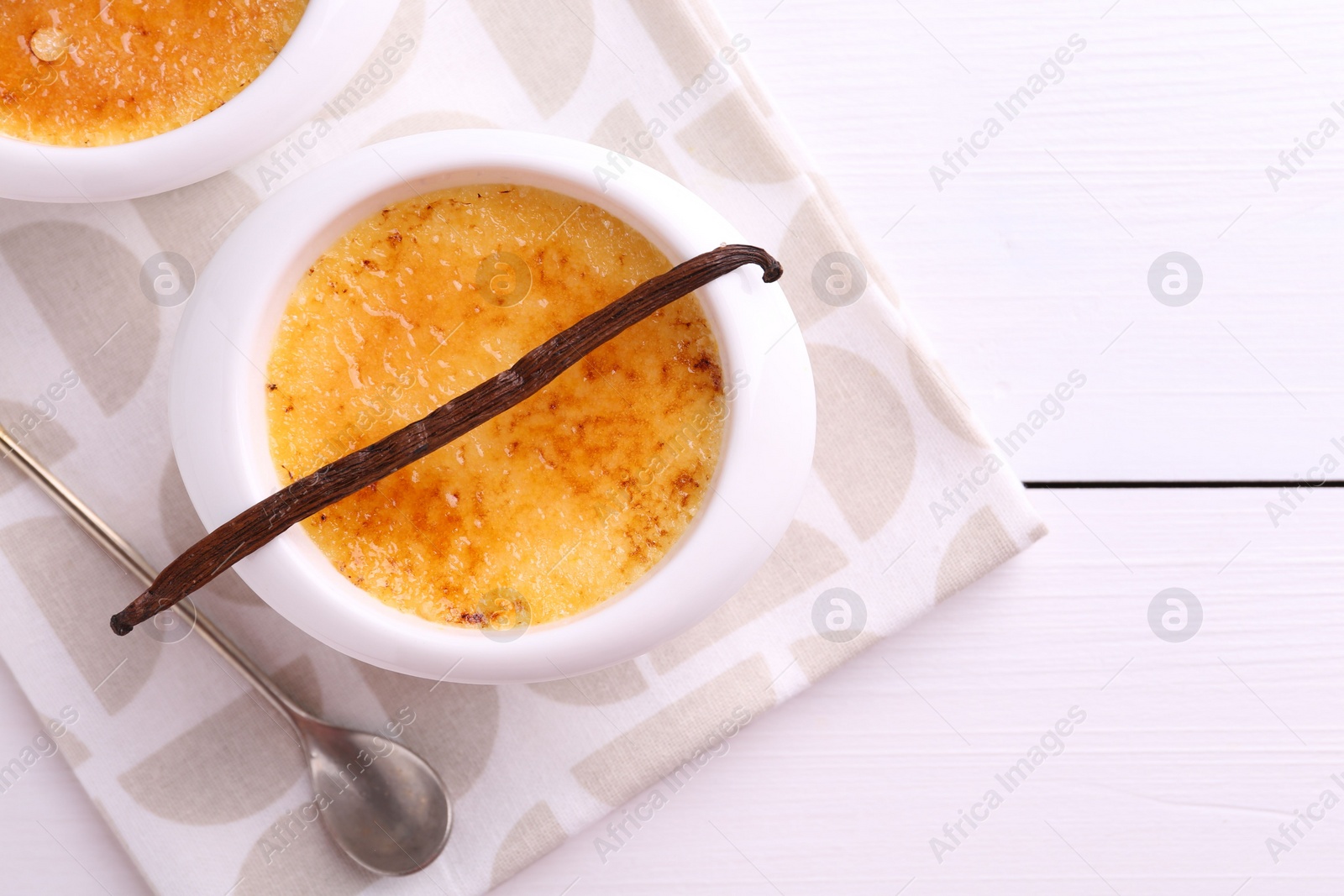 Photo of Delicious creme brulee in bowls, vanilla pod and spoon on white wooden table, flat lay