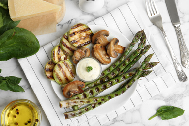 Tasty grilled asparagus served with sauce, zucchini and mushrooms on white marble table, flat lay