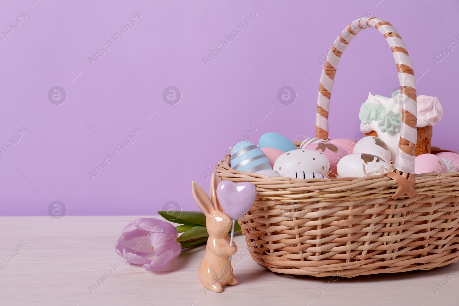 Photo of Easter basket with painted eggs, cake, flower and rabbit figure on white wooden table. Space for text