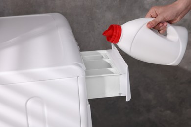 Photo of Woman pouring fabric softener from bottle into washing machine near grey wall, closeup