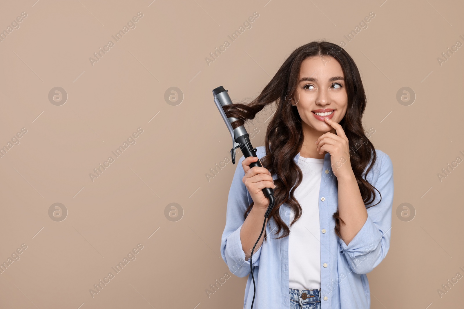 Photo of Happy woman using curling hair iron on beige background. Space for text