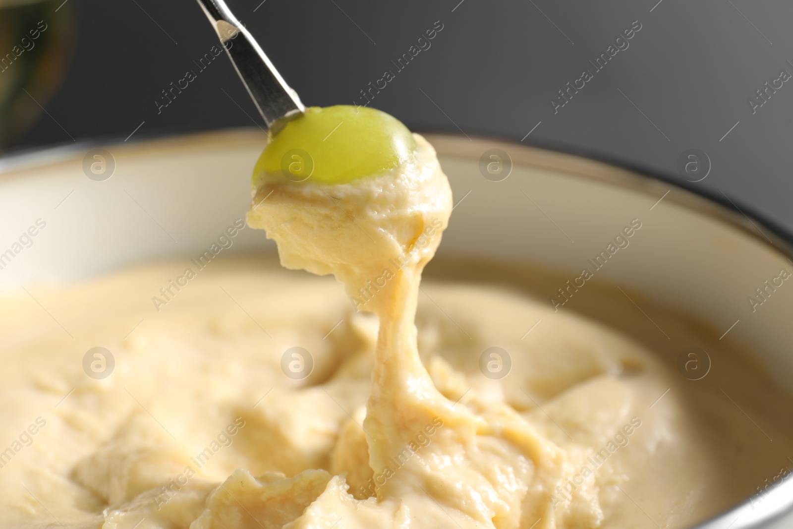 Photo of Dipping piece of grape into fondue pot with melted cheese on grey background, closeup