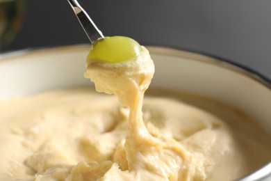 Dipping piece of grape into fondue pot with melted cheese on grey background, closeup