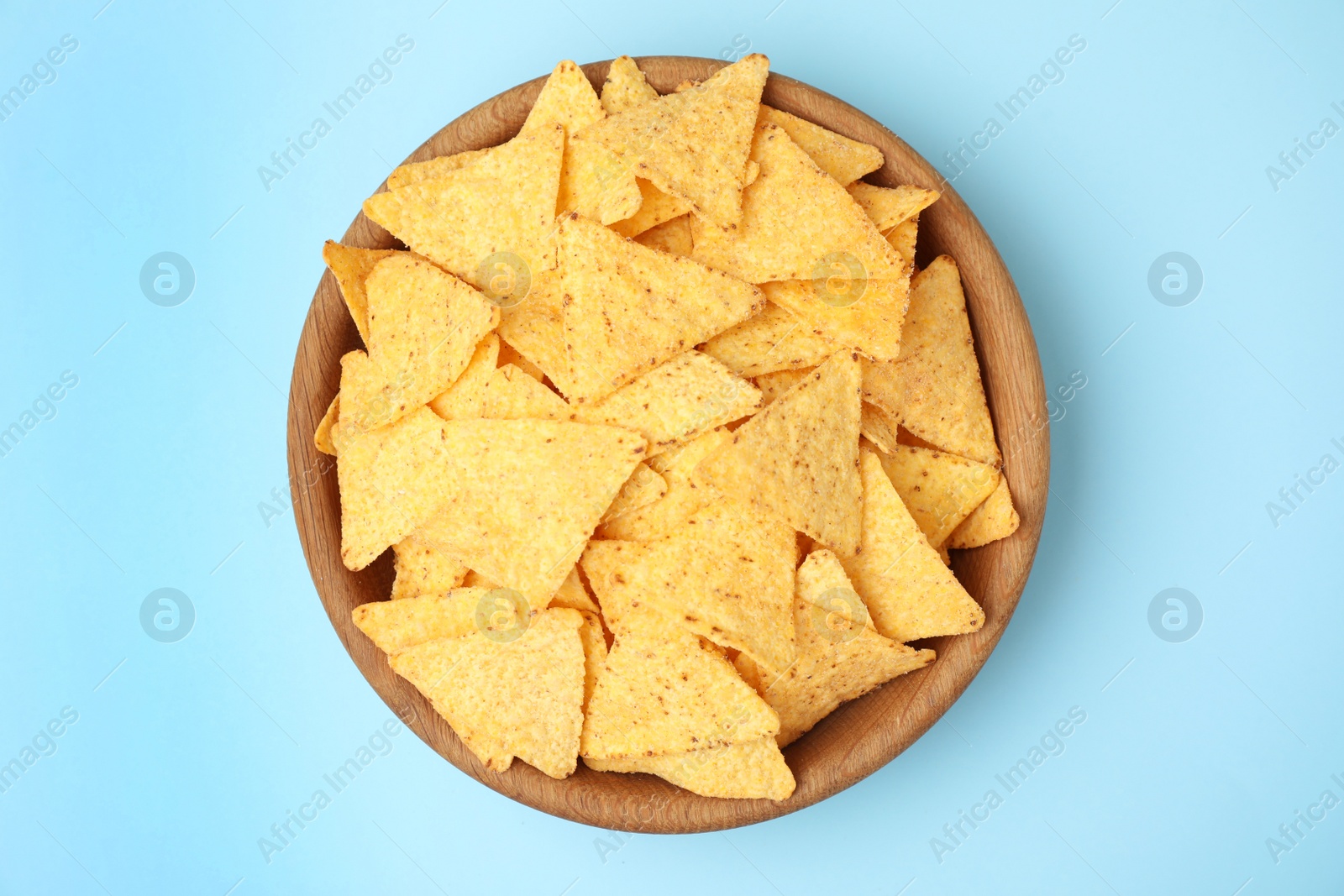 Photo of Wooden bowl of tasty Mexican nachos chips on light blue background, top view