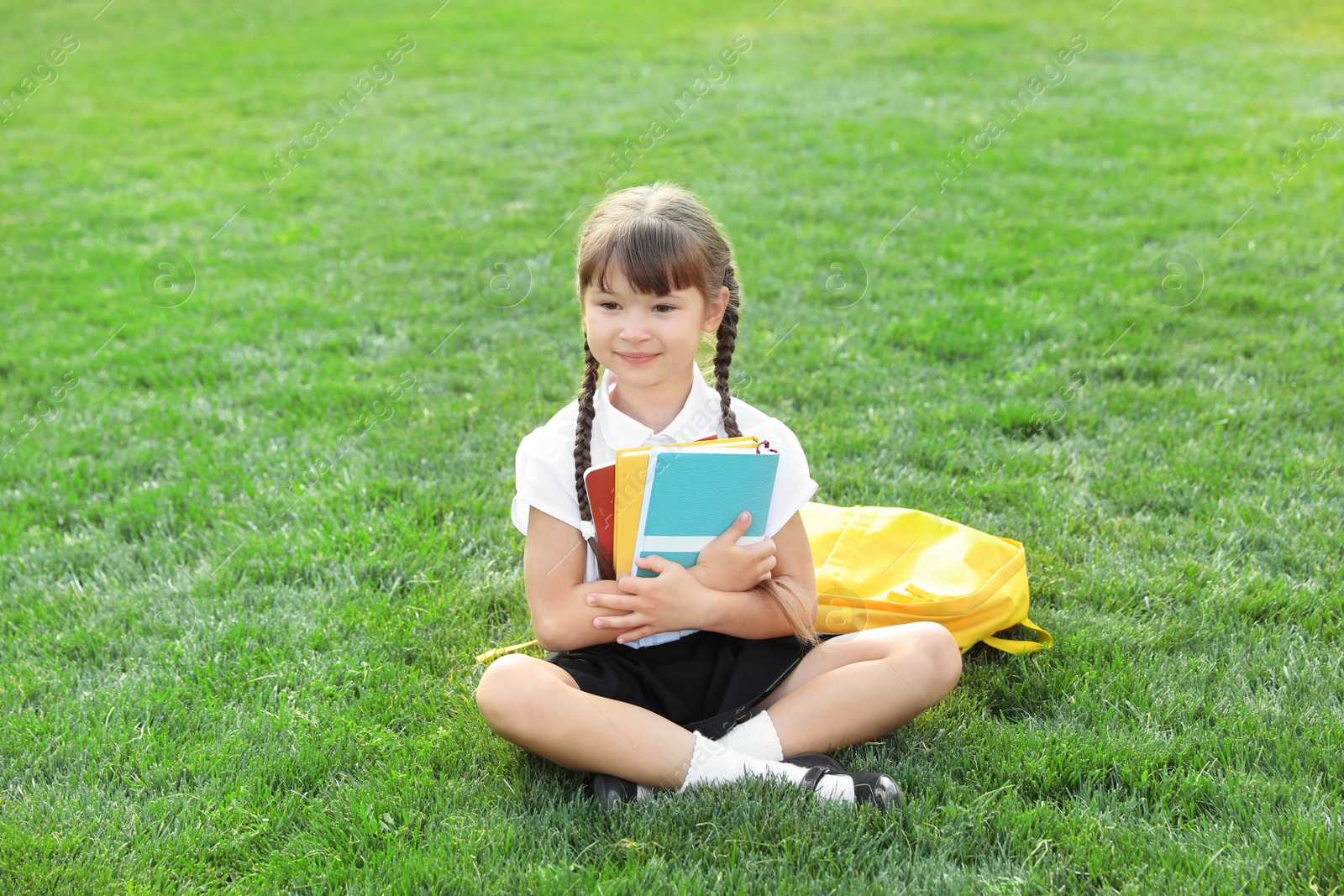 Photo of Cute girl with school stationery sitting on green lawn outdoors