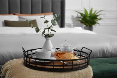 Photo of Wicker tray with vase of eucalyptus leaves, tea and cookies on ottoman in bedroom. Interior design