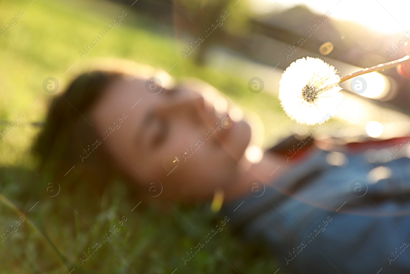 Photo of Young woman lying on green grass in park, focus on dandelion. Allergy free concept