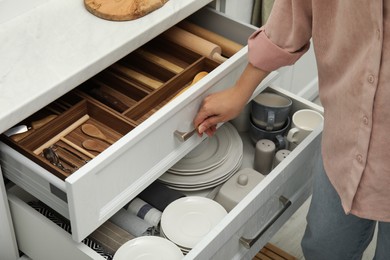 Photo of Woman opening drawers of kitchen cabinet with different dishware and utensils, closeup