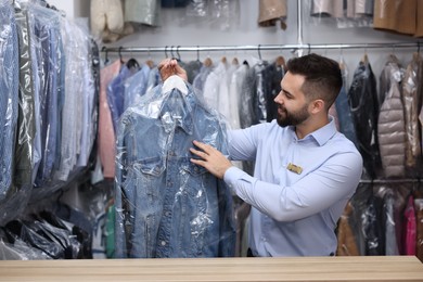 Photo of Dry-cleaning service. Happy worker holding hanger with denim jacket in plastic bag at counter indoors