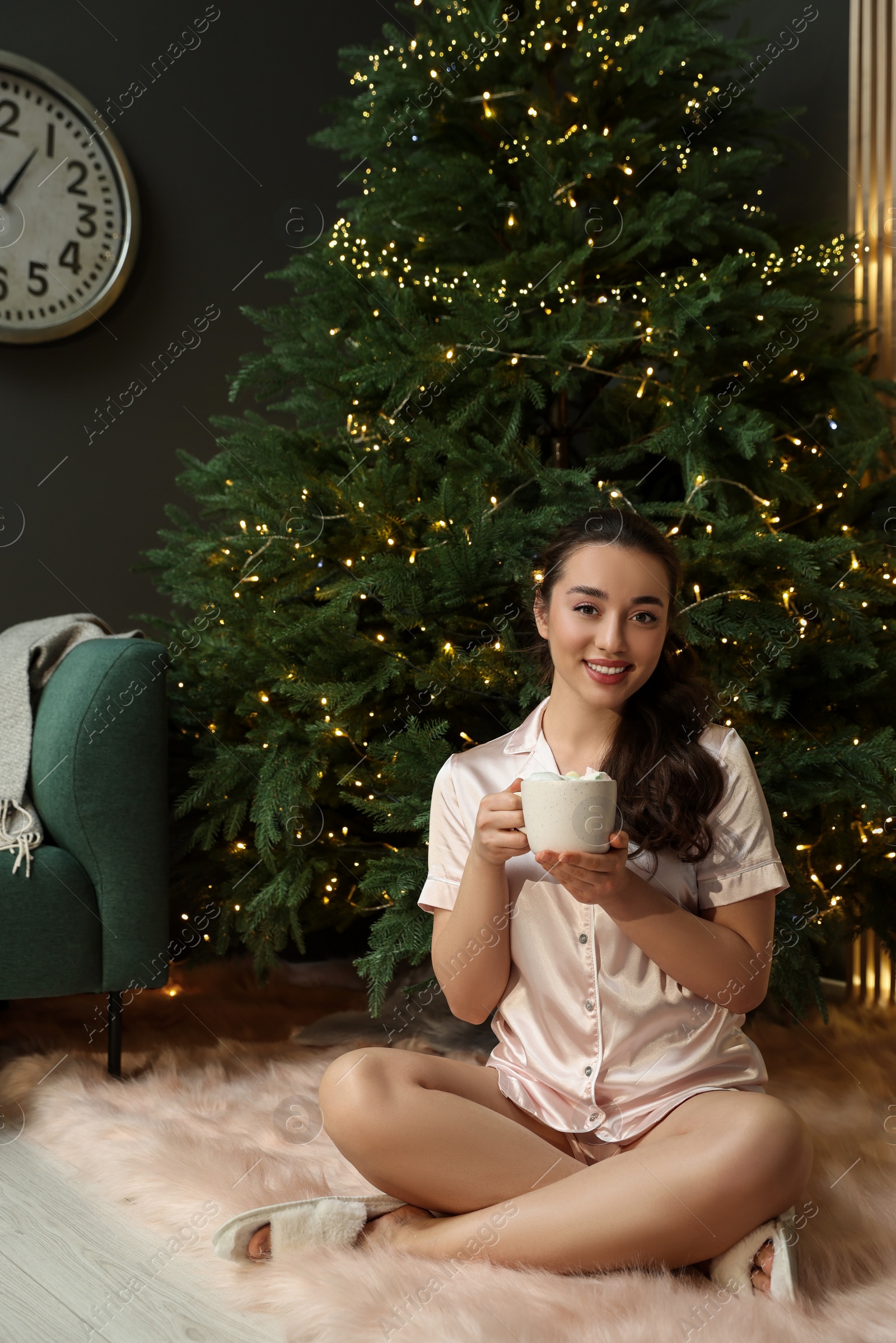 Photo of Beautiful woman with cup of tasty drink near Christmas tree at home