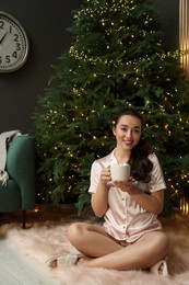 Photo of Beautiful woman with cup of tasty drink near Christmas tree at home