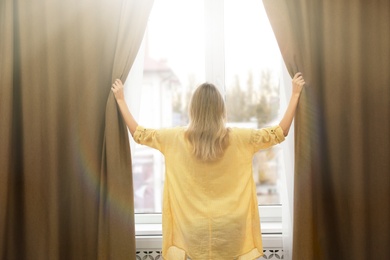 Young woman opening window curtains at home, back view