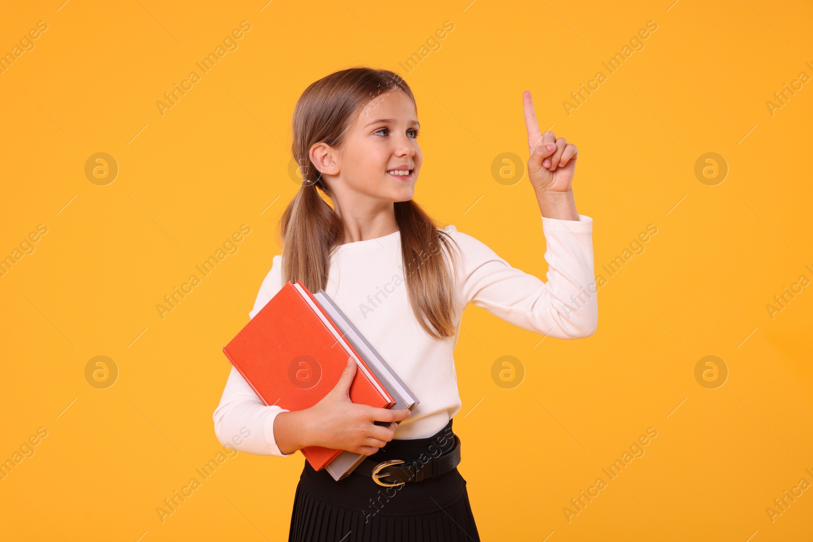 Photo of Happy schoolgirl with books pointing at something on orange background