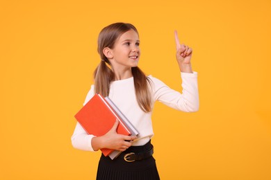 Happy schoolgirl with books pointing at something on orange background