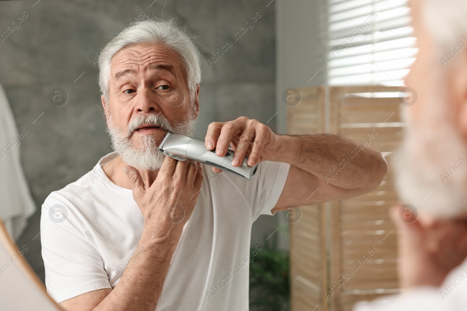 Photo of Senior man trimming beard near mirror in bathroom