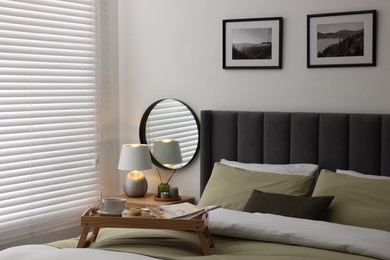 Wooden tray table with cup of drink and magazines on bed near window with horizontal blinds in room