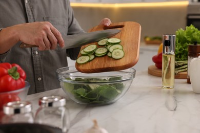 Photo of Cooking process. Man adding cut cucumber into bowl at white marble countertop in kitchen, closeup