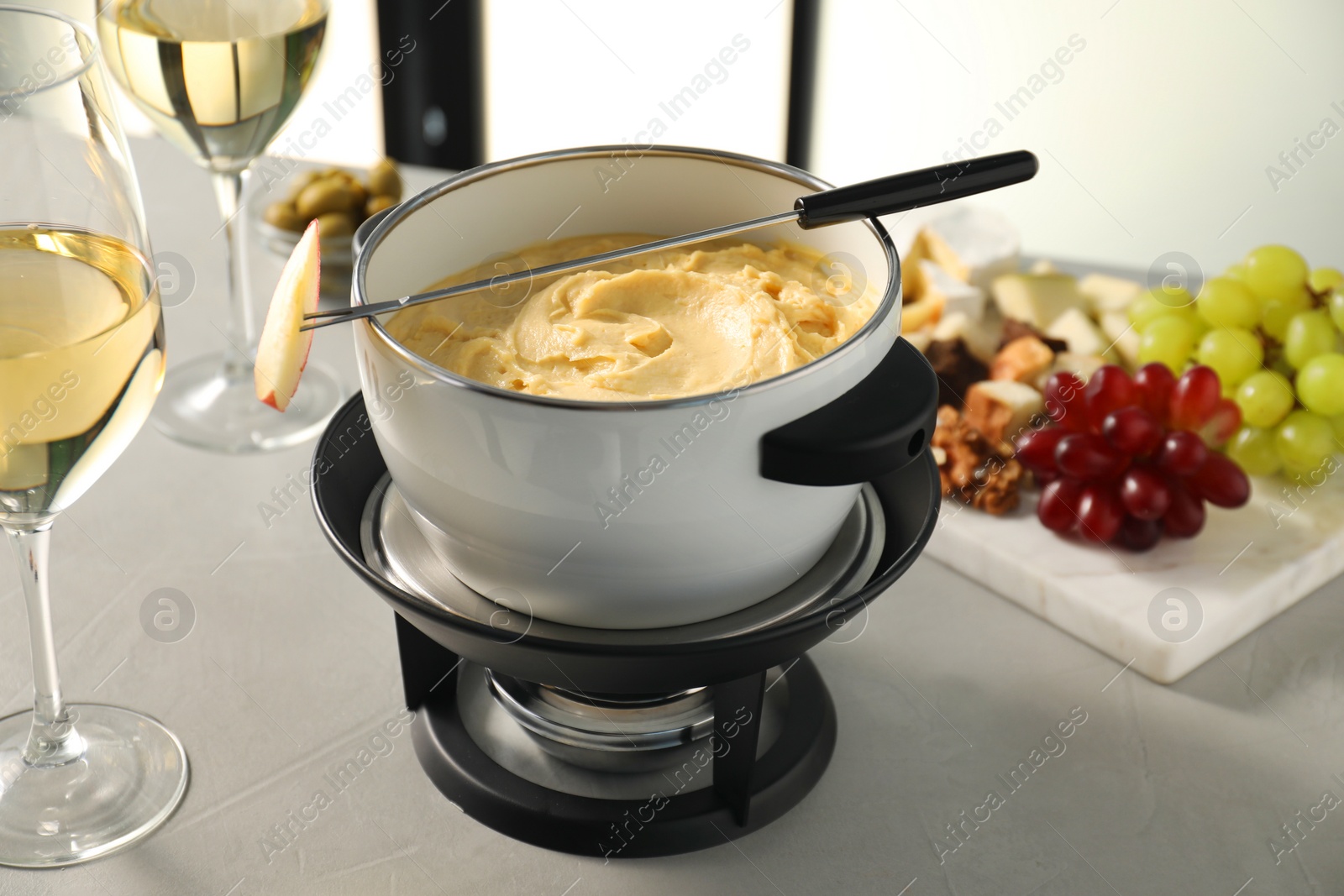 Photo of Fork with piece of apple, melted cheese in fondue pot, wine and products on grey table, closeup