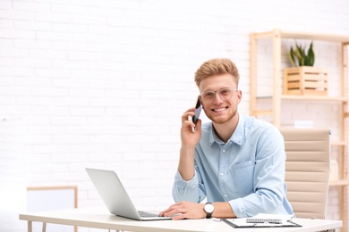 Photo of Handsome young man talking on phone while working with laptop at table in office