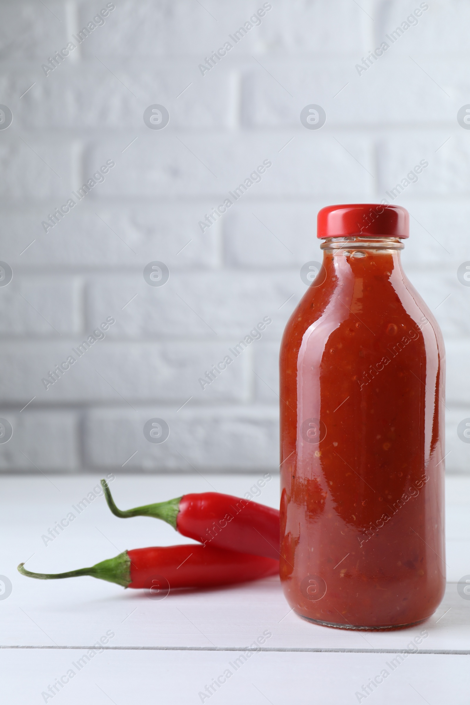 Photo of Spicy chili sauce in bottle and peppers on white wooden table, space for text