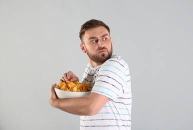 Greedy young man hiding bowl with chips on light grey background