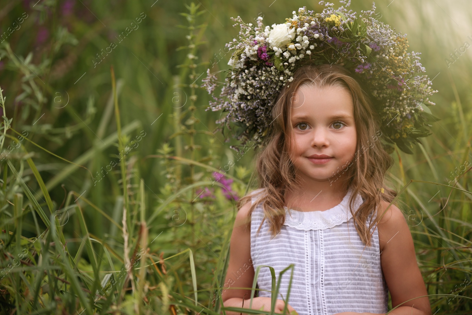 Photo of Cute little girl wearing wreath made of beautiful flowers in field