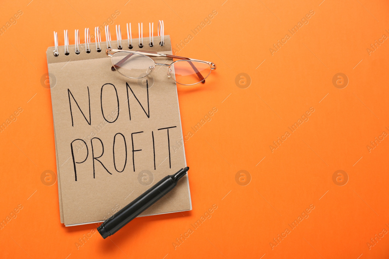 Photo of Notebook with phrase Non Profit, glasses and pen on orange background, flat lay. Space for text