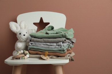 Stack of baby clothes, pacifier and toys on chair near light brown wall. Space for text