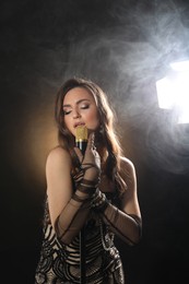 Photo of Beautiful young woman with microphone singing on dark background with smoke