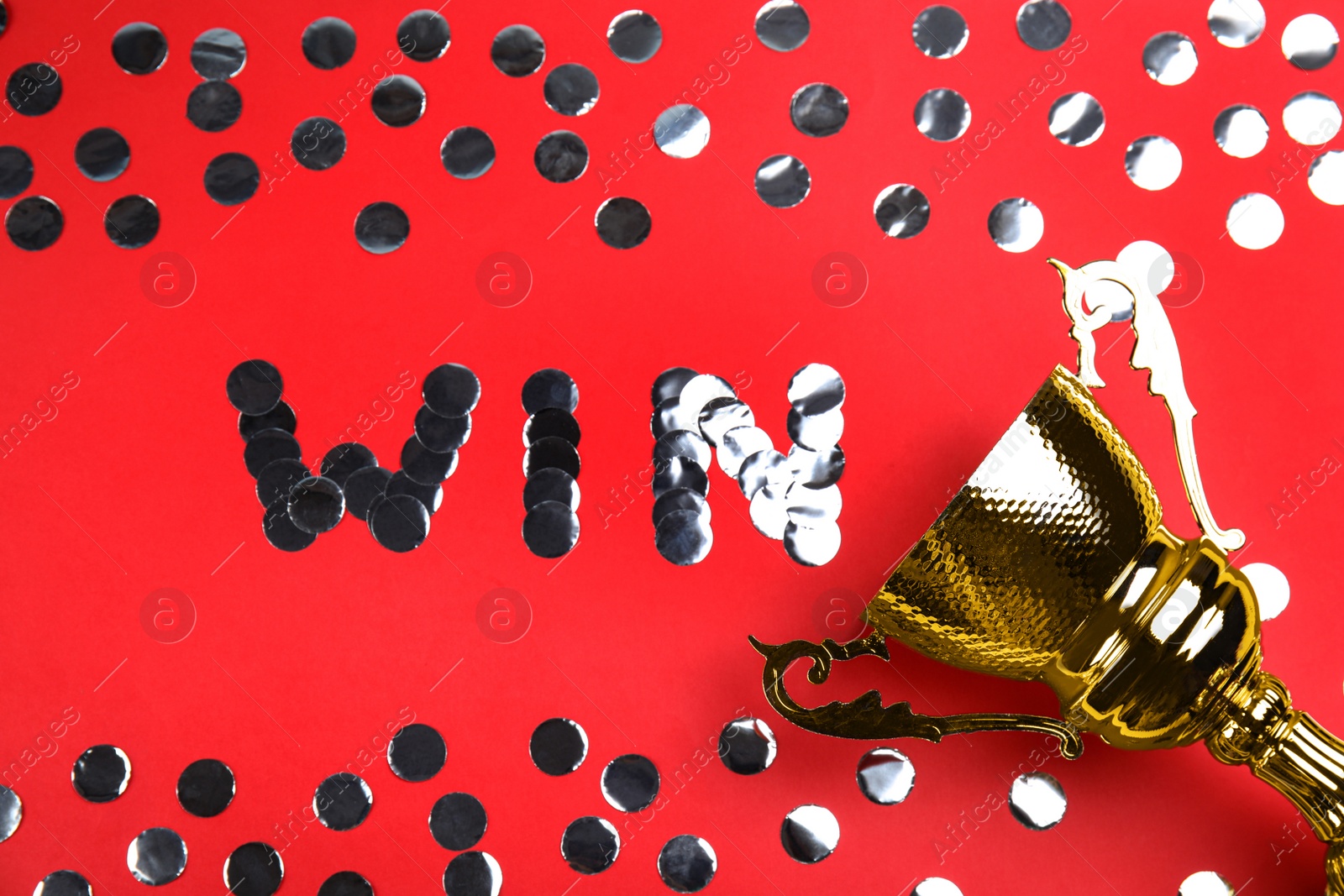 Photo of Gold trophy cup and word WIN made with silver confetti on red background, flat lay