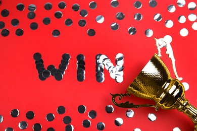 Photo of Gold trophy cup and word WIN made with silver confetti on red background, flat lay